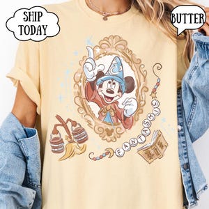 K&ouml;nnte beinhalten: Hellgelbes T-Shirt mit einem Cartoon-Bild von Mickey Mouse als Zauberer in einem dekorativen Rahmen. Das Wort "FANTASTIC" ist in einem Perlenketten-Design geschrieben. Zus&auml;tzliche Grafiken sind ein Besen und ein Buch.