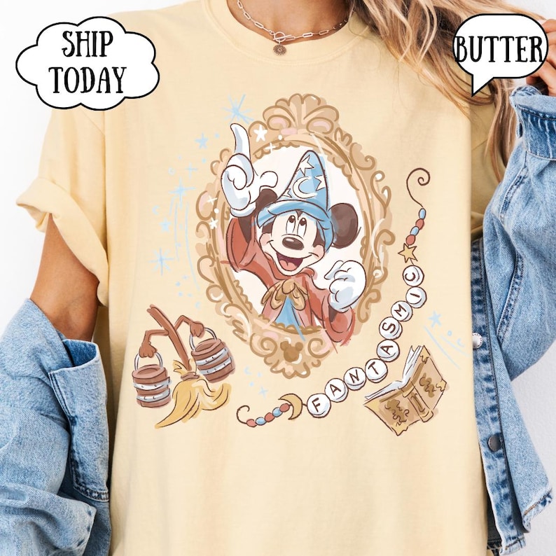 K&ouml;nnte beinhalten: Hellgelbes T-Shirt mit einem Cartoon-Bild von Mickey Mouse als Zauberer in einem dekorativen Rahmen. Das Wort "FANTASTIC" ist in einem Perlenketten-Design geschrieben. Zus&auml;tzliche Grafiken sind ein Besen und ein Buch.