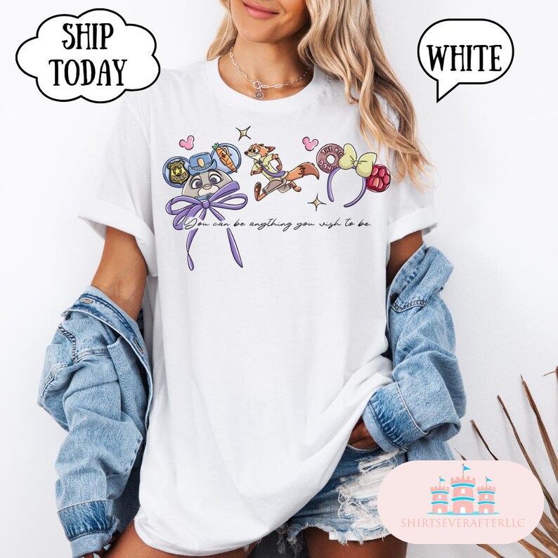 May include: White t-shirt featuring a colorful graphic of cartoon characters and headbands. The text "You can be anything you wish to be" is printed below the graphic. The shirt has short sleeves.
