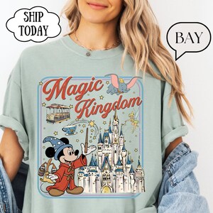 May include: A light green t-shirt featuring a colorful graphic of Mickey Mouse as a wizard, a castle, and the words "Magic Kingdom." The design includes images of Dumbo, a trolley car, and Tinkerbell. The shirt has a relaxed fit.