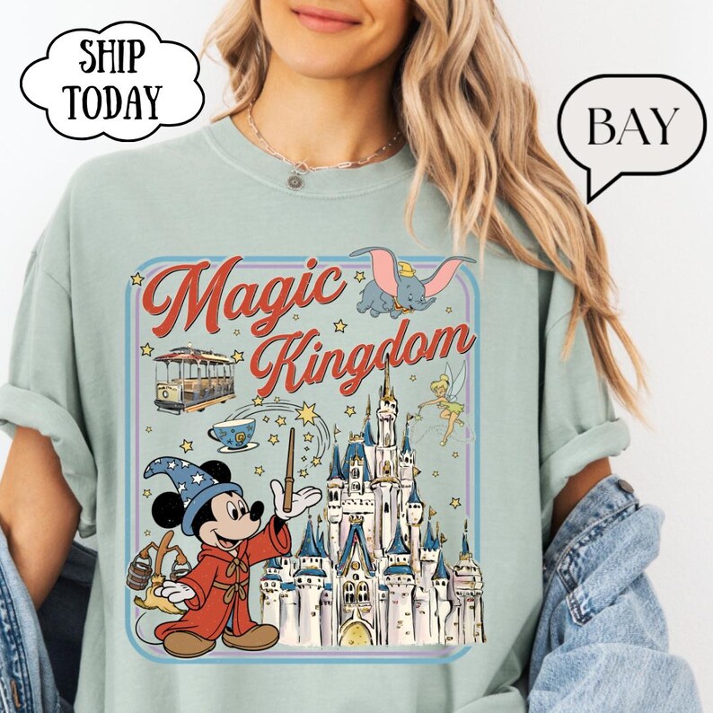 May include: A light green t-shirt featuring a colorful graphic of Mickey Mouse as a wizard, a castle, and the words "Magic Kingdom." The design includes images of Dumbo, a trolley car, and Tinkerbell. The shirt has a relaxed fit.