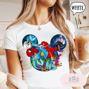 Puede incluir: Camiseta blanca con un gr&aacute;fico colorido de los personajes de La Sirenita de Disney dentro de un dise&ntilde;o de oreja de Mickey Mouse. El dise&ntilde;o incluye a Ariel, &Uacute;rsula y otros personajes. La palabra "WHITE" est&aacute; en un bocadillo.