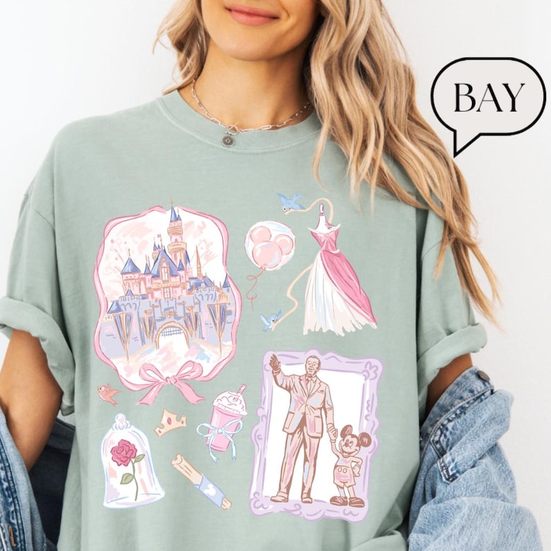 Op de afbeelding: Saliegroen T-shirt met diverse illustraties, waaronder een roze kasteel, een roze en witte jurk en een ingelijste afbeelding van een man en een kind. Andere afbeeldingen zijn een roos, een kroon en een drankje.
