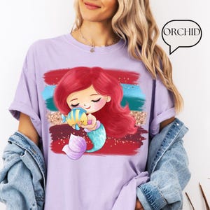 Puede incluir: Camiseta lila claro con una sirena de dibujos animados con pelo rojo y un pez azul y amarillo. La sirena abraza al pez. El fondo tiene pinceladas rojas, azules y doradas. La palabra "ORCHID" est&aacute; en un bocadillo.