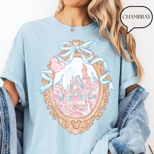 Puede incluir: Camiseta azul claro con una ilustraci&oacute;n enmarcada en oro de un castillo de cuento de hadas con detalles en rosa y azul. Una cinta azul claro y globos rosas adornan la parte superior del marco. La palabra "CHAMBRAY" est&aacute; en una burbuja.