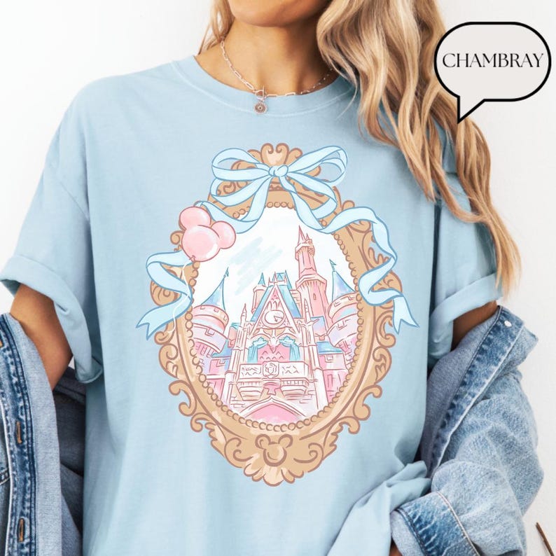 Puede incluir: Camiseta azul claro con una ilustraci&oacute;n enmarcada en oro de un castillo de cuento de hadas con detalles en rosa y azul. Una cinta azul claro y globos rosas adornan la parte superior del marco. La palabra "CHAMBRAY" est&aacute; en una burbuja.