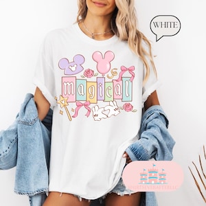 Op de afbeelding: Wit t-shirt met het woord "magical" in pastelkleuren, met cartoonafbeeldingen van een muizenkop, ballon, strik, roos, konijn en een toverstaf. Het woord "WHITE" staat in een tekstballon.