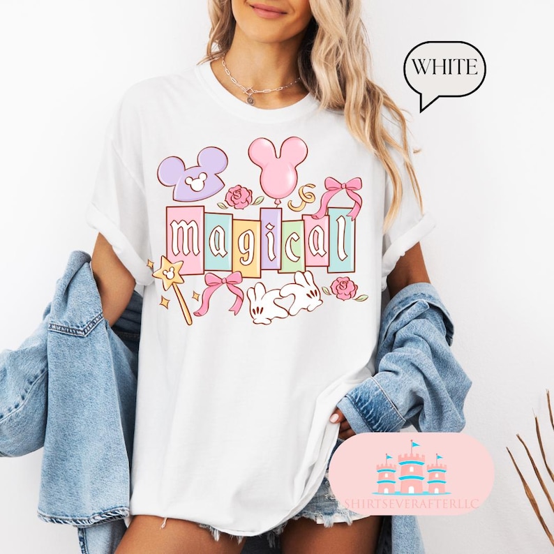 Op de afbeelding: Wit t-shirt met het woord "magical" in pastelkleuren, met cartoonafbeeldingen van een muizenkop, ballon, strik, roos, konijn en een toverstaf. Het woord "WHITE" staat in een tekstballon.