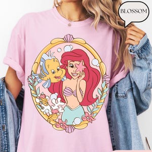 May include: Pink t-shirt featuring a cartoon illustration of Ariel from The Little Mermaid, with Flounder. The design is framed in a gold oval with floral and seashell accents. The word "BLOSSOM" is in a speech bubble.
