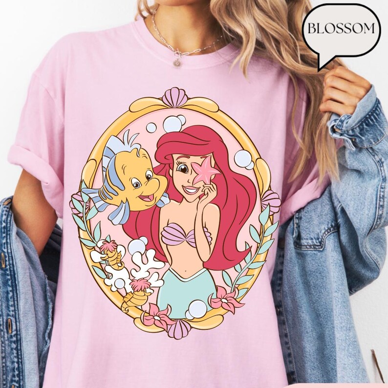 May include: Pink t-shirt featuring a cartoon illustration of Ariel from The Little Mermaid, with Flounder. The design is framed in a gold oval with floral and seashell accents. The word "BLOSSOM" is in a speech bubble.