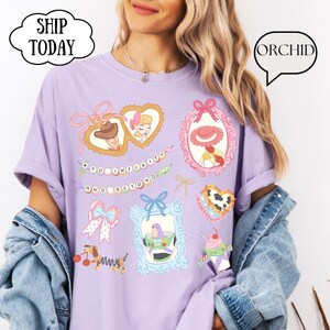 Op de afbeelding: Een lichtpaars T-shirt met illustraties van stripfiguren in kaders en hartvormen. Het shirt heeft de tekst "TO INFINITY AND BEYOND". De mouwen zijn opgerold en het wordt gedragen met een spijkerjasje.
