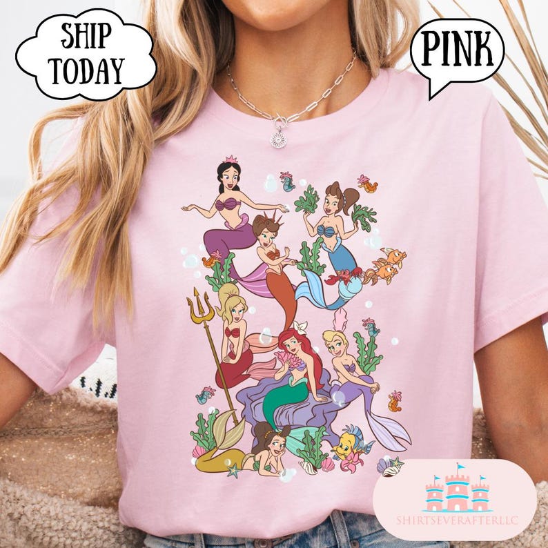 Op de afbeelding: Zachtroze T-shirt met een kleurrijke illustratie van verschillende zeemeerminnen met verschillende haar- en staartkleuren. Het shirt heeft het woord "PINK" in een tekstballon. Het shirt heeft ook de tekst "SHIP TODAY" in een tekstballon.