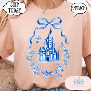 以下が含まれることがあります： ピーチ色のTシャツ。青い水彩画で城、鳥、花の要素が描かれています。デザインにはリボンと「H/PEACH」の文字が含まれています。Tシャツは丸いネックラインです。