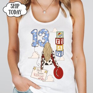 May include: A white tank top featuring a graphic of a person with long blonde hair wearing a cow-print jacket with red fringe. The design includes a blue cloud-patterned number 13 balloon, stacked alphabet blocks, and a sign reading Tays Room Keep Out.