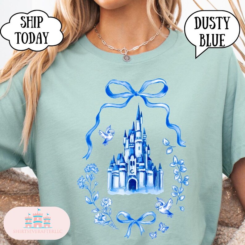 以下が含まれることがあります： 青みがかった水色のTシャツ。青い花のリースの中に城の水彩画イラストが描かれており、リボンと鳥がアクセントになっています。「Dusty Blue」と「Ship Today」の文字が吹き出しに入っています。
