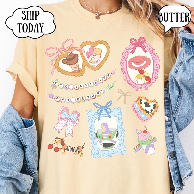 Op de afbeelding: Boterkleurig T-shirt met cartoonkarakterafbeeldingen in roze, blauw en geel. Het shirt bevat ingelijste afbeeldingen van personages, strikken en de zin "TO INFINITY AND BEYOND". De woorden "SHIP TODAY" en "BUTTER" zijn ook zichtbaar.