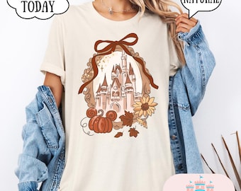 May include: Cream-colored t-shirt featuring a fall-themed graphic. The design includes a castle, pumpkin, sunflower, and leaves within a decorative frame. The shirt also has the text "SHIP TODAY" and "NATURAL".