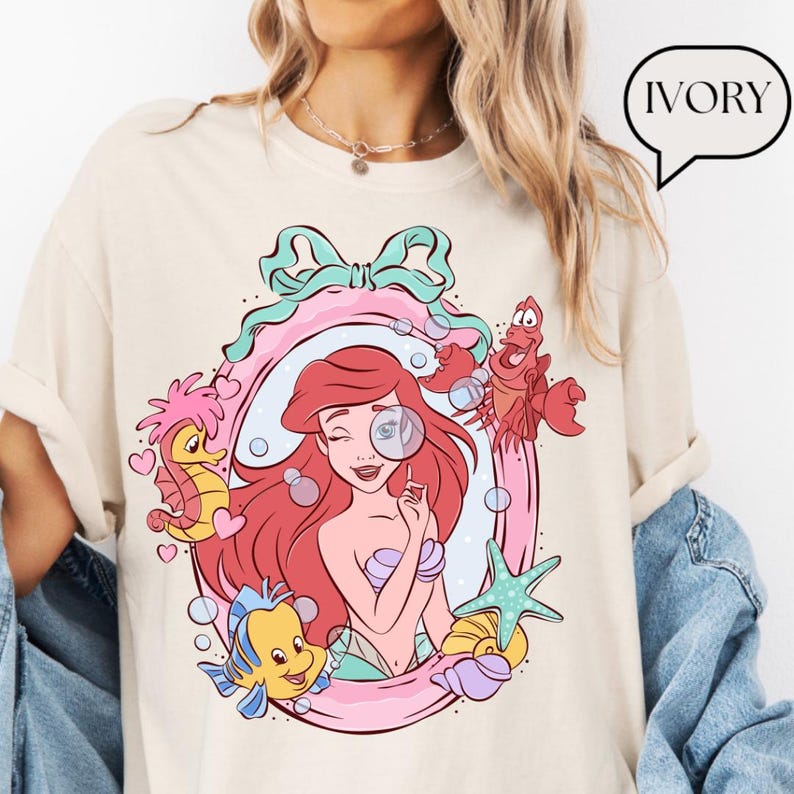 May include: Ivory-colored t-shirt featuring a colorful illustration of Ariel from The Little Mermaid. The design includes Ariel, Flounder, Sebastian, a seahorse, and a starfish, all within a pink and blue frame. The word "IVORY" is in a speech bubble.
