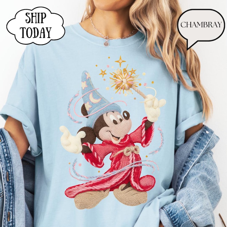 May include: Pale blue t-shirt with an embroidered Mickey Mouse design, dressed as a sorcerer with a wand and starburst. The shirt has rolled sleeves and the text "SHIP TODAY" in a cloud-shaped speech bubble. The word "CHAMBRAY" is also visible.