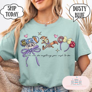 May include: Dusty blue t-shirt featuring cartoon characters and the text "You can be anything you wish to be." The design includes a police badge, a fox, a donut, and a bow. The shirt has a crew neck and short sleeves.