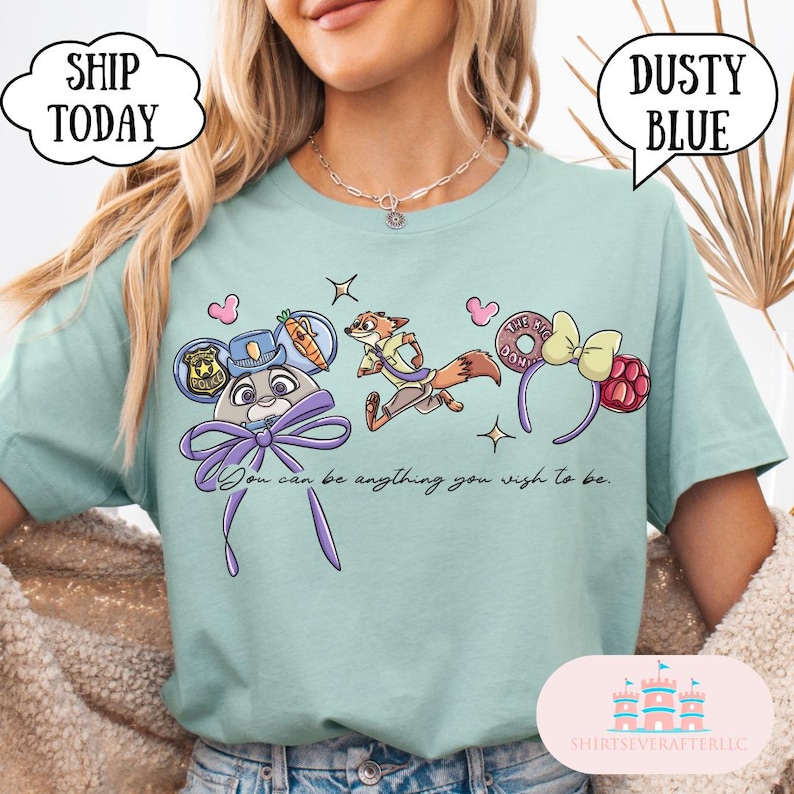 May include: Dusty blue t-shirt featuring cartoon characters and the text "You can be anything you wish to be." The design includes a police badge, a fox, a donut, and a bow. The shirt has a crew neck and short sleeves.