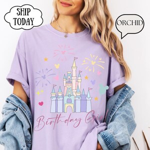 May include: A lilac t-shirt with a castle graphic, fireworks, and stars. The words "Birthday Girl" are in cursive. The shirt also has the text "SHIP TODAY" and "ORCHID" in speech bubbles.