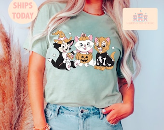 以下が含まれることがあります： 薄い緑色のTシャツに、ハロウィンのコスチュームを着た3匹の漫画の猫のグラフィックが描かれています。猫たちは、カボチャ、ロリポップ、骨を持っています。Tシャツには「トリックオアトリート」の文字が書かれています。