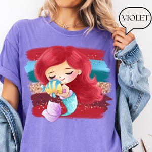 Puede incluir: Camiseta violeta con una sirena de dibujos animados abrazando a un pez amarillo y azul. La sirena tiene el pelo rojo y una cola turquesa. El dise&ntilde;o incluye pinceladas rojas, azules y doradas. La palabra "VIOLET" est&aacute; en un bocadillo.