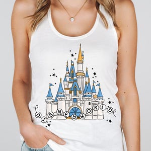 May include: White tank top featuring a colorful illustration of a fairytale castle with blue turrets and gold accents. The words "Disney World" are written in a circular design around the castle, with black stars scattered around the image.