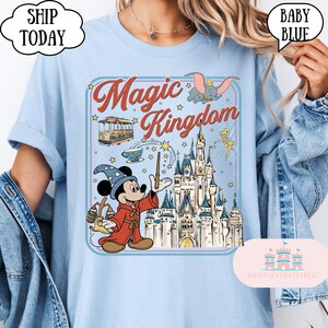 May include: Baby blue t-shirt featuring a colorful graphic of Mickey Mouse as a sorcerer, a castle, and the words "Magic Kingdom". The design includes a cable car, Dumbo, and Tinkerbell. The shirt has a crew neck.