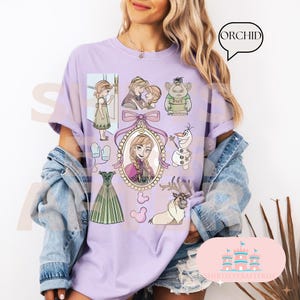 Könnte beinhalten: Ein helllila T-Shirt mit Cartoon-Illustrationen aus dem Film Frozen. Das Design umfasst Charaktere wie Anna, Elsa, Olaf und Sven sowie andere verwandte Elemente. Das Wort "ORCHID" befindet sich in einer Sprechblase.