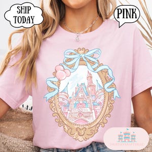 Puede incluir: Camiseta rosa claro con una ilustraci&oacute;n enmarcada en oro de un castillo de cuento de hadas con detalles azules y globos rosas. La camiseta tiene la palabra "PINK" en un bocadillo.