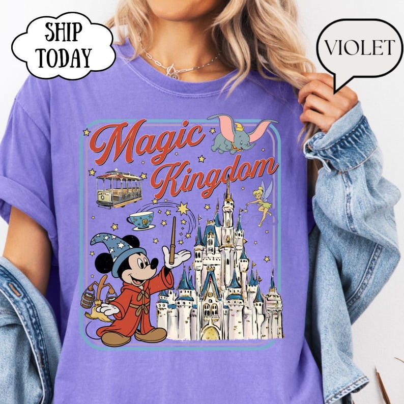 May include: Violet-colored t-shirt featuring a graphic of Mickey Mouse in a wizard's robe, a castle, and the words "Magic Kingdom." The design includes Dumbo, Tinkerbell, and a trolley car. The shirt has a relaxed fit.