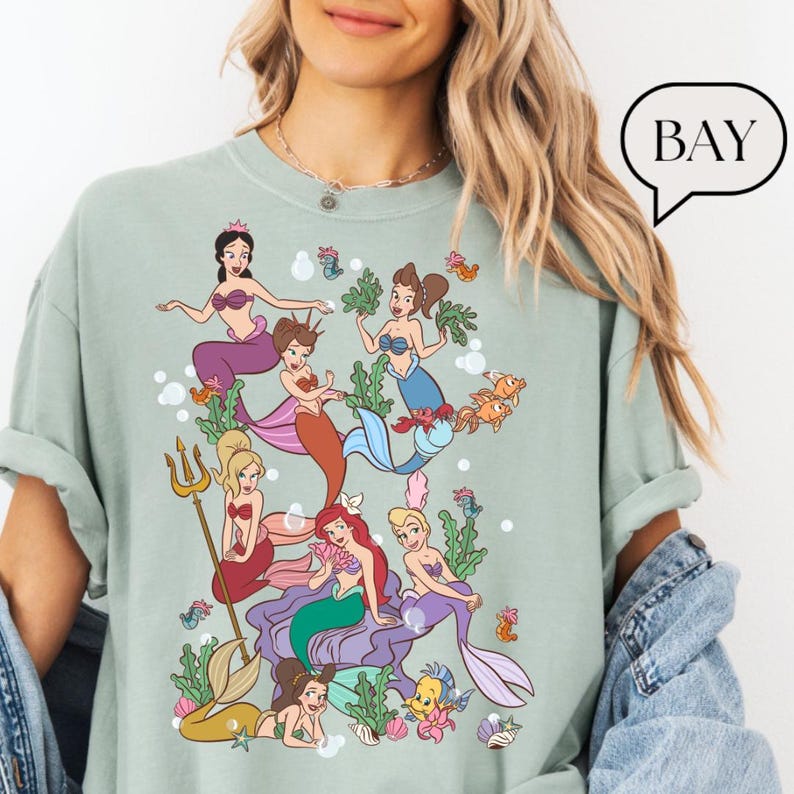 Op de afbeelding: Lichtgroen T-shirt met een kleurrijke illustratie van zeemeerminnen. Het ontwerp toont diverse zeemeerminnen met verschillende haarkleuren en staarten, omringd door waterplanten en vissen. Het woord "BAY" staat in een tekstballon.