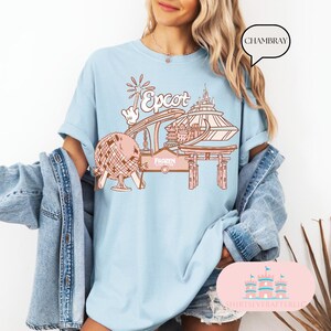 May include: A light blue t-shirt featuring a graphic design of Epcot, a Disney theme park. The design includes a pink and white Spaceship Earth, a pink and white Test Track ride, a pink and white Frozen Ever After ride, and a pink and white World Showcase entrance. The text "Epcot" is in pink.