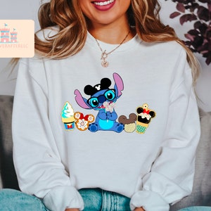 Puede incluir: Una sudadera blanca con un personaje azul Stitch que lleva orejas de Mickey Mouse y gafas de sol. Stitch sostiene un cono de helado de Mickey Mouse y est&aacute; rodeado de otras golosinas de Disney.