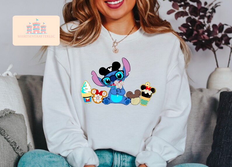 Puede incluir: Una sudadera blanca con un personaje azul Stitch que lleva orejas de Mickey Mouse y gafas de sol. Stitch sostiene un cono de helado de Mickey Mouse y est&aacute; rodeado de otras golosinas de Disney.
