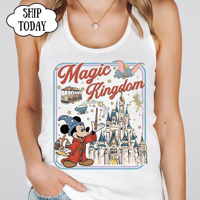 May include: White tank top featuring a colorful graphic of Mickey Mouse as a sorcerer, a castle, and the words "Magic Kingdom." The design includes Dumbo, a trolley car, and Tinkerbell. The top has a round neckline.