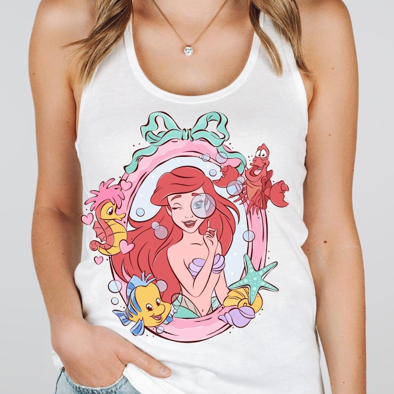 May include: White tank top featuring a cartoon illustration of Ariel from The Little Mermaid. The design includes Ariel, Flounder, Sebastian, and other sea creatures within a pink frame with a bow.