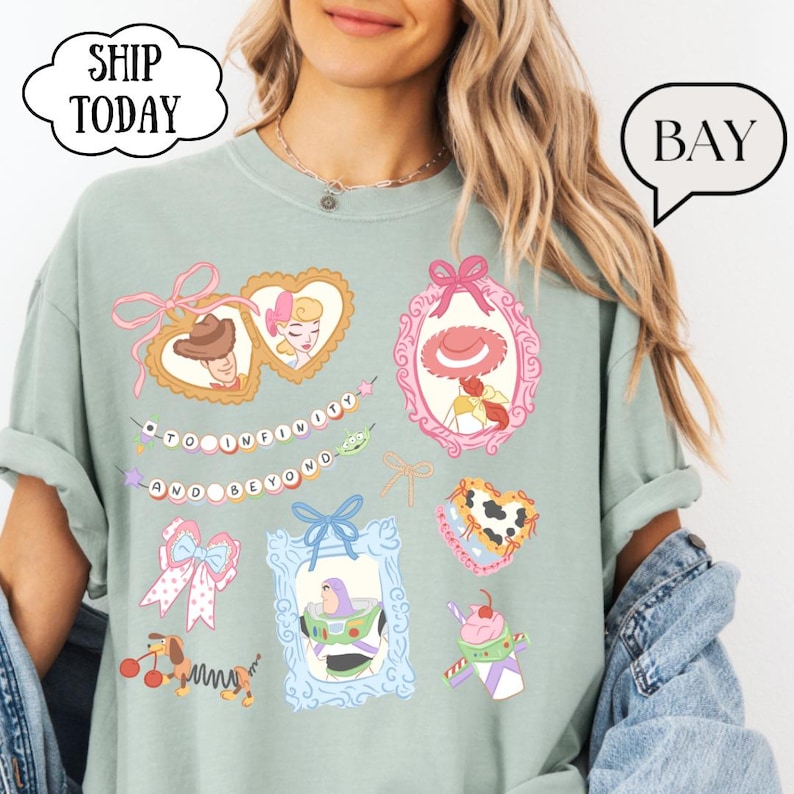 Op de afbeelding: Saliegroen T-shirt met kleurrijke cartoonafbeeldingen van personages en elementen uit een populaire animatiefilm. Het shirt bevat de zin "TO INFINITY AND BEYOND" en diverse ingelijste afbeeldingen. Een tekstballon in de vorm van een wolk zegt "SHIP TODAY".