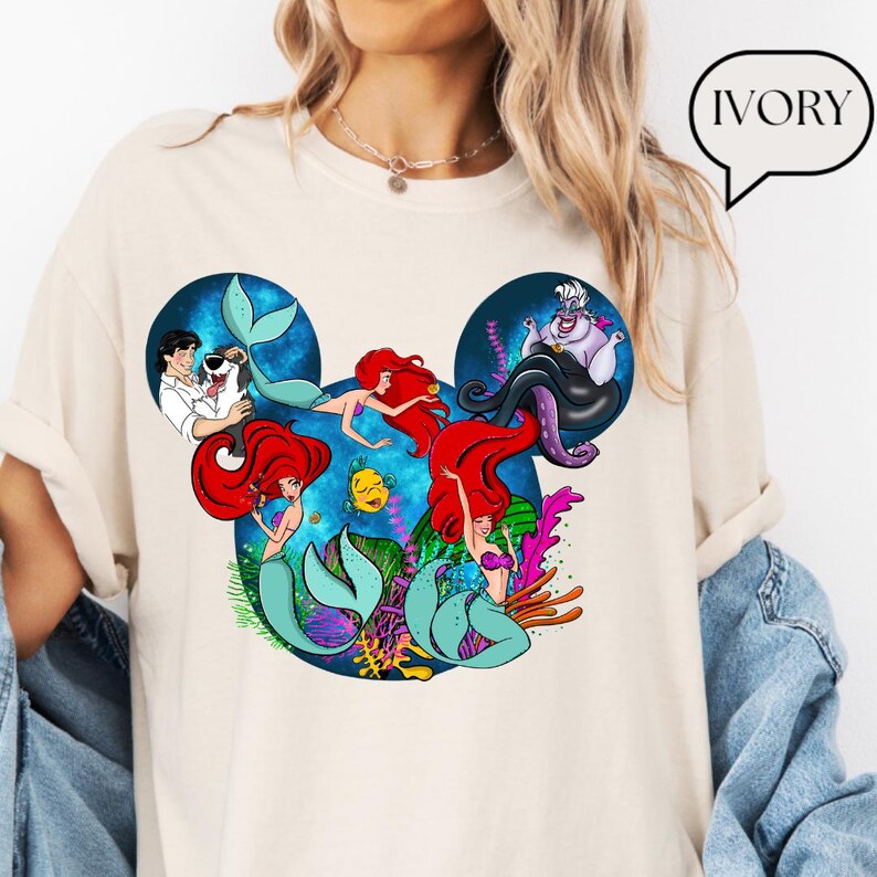 Puede incluir: Camiseta de color marfil con un gr&aacute;fico de los personajes de La Sirenita de Disney dentro de la forma de la cabeza de Mickey Mouse. El dise&ntilde;o incluye a Ariel, &Uacute;rsula, el Pr&iacute;ncipe Eric y Flounder, con colores vibrantes e ilustraciones detalladas.