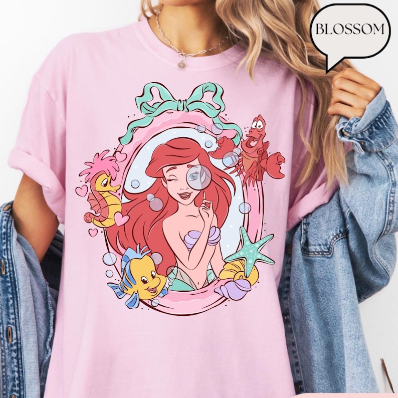 May include: Pink t-shirt featuring a cartoon illustration of a red-haired mermaid with a magnifying glass, surrounded by sea creatures. The design includes a seahorse, a fish, a crab, and a starfish, all within a pink frame. The word "BLOSSOM" is in a speech bubble.