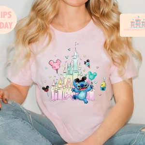 May include: A pink t-shirt with a colorful illustration of Stitch from Lilo & Stitch in front of a pastel-colored version of Cinderella's Castle. The illustration includes Mickey Mouse ears, a cotton candy, a Mickey Mouse ice cream cone, and a cupcake.