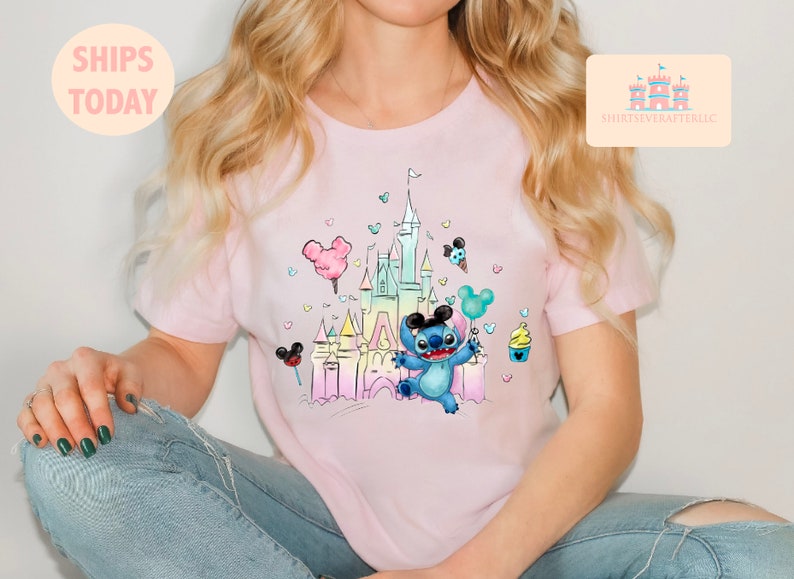 May include: A pink t-shirt with a colorful illustration of Stitch from Lilo & Stitch in front of a pastel-colored version of Cinderella's Castle. The illustration includes Mickey Mouse ears, a cotton candy, a Mickey Mouse ice cream cone, and a cupcake.