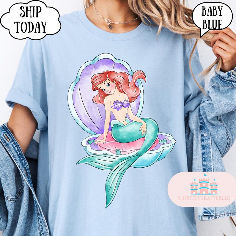 May include: Pale blue t-shirt with a watercolour illustration of a mermaid in a shell. The mermaid has red hair, a purple seashell bra, and a teal tail. The shirt has the text "Ship Today" and "Baby Blue".