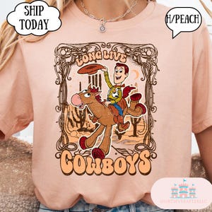 May include: Peach-colored t-shirt featuring a cartoon cowboy riding a horse, with the words "Long Live Cowboys" in a decorative frame. The design includes a cowboy with a hat and a desert landscape.