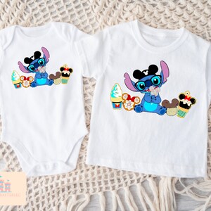 Puede incluir: Dos camisetas blancas con un personaje de dibujos animados azul que lleva orejas de Mickey Mouse y gafas de sol. El personaje sostiene un cono de helado de Mickey Mouse y est&aacute; rodeado de otras golosinas de Disney. Una camiseta es un body y la otra es una camiseta de manga corta.