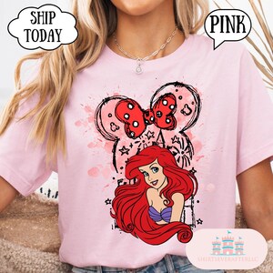 May include: A light pink t-shirt featuring a graphic of Ariel from The Little Mermaid with red hair and a purple top. The design includes a red polka dot bow and a Mickey Mouse head outline. The shirt has the word "PINK" in a speech bubble.