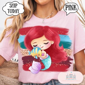 Puede incluir: Camiseta rosa claro con una ilustraci&oacute;n de dibujos animados de una sirena con cabello rojo y cola turquesa, sosteniendo un pez amarillo y azul. El fondo tiene pinceladas rojas, azules y doradas. Globo de texto "PINK".
