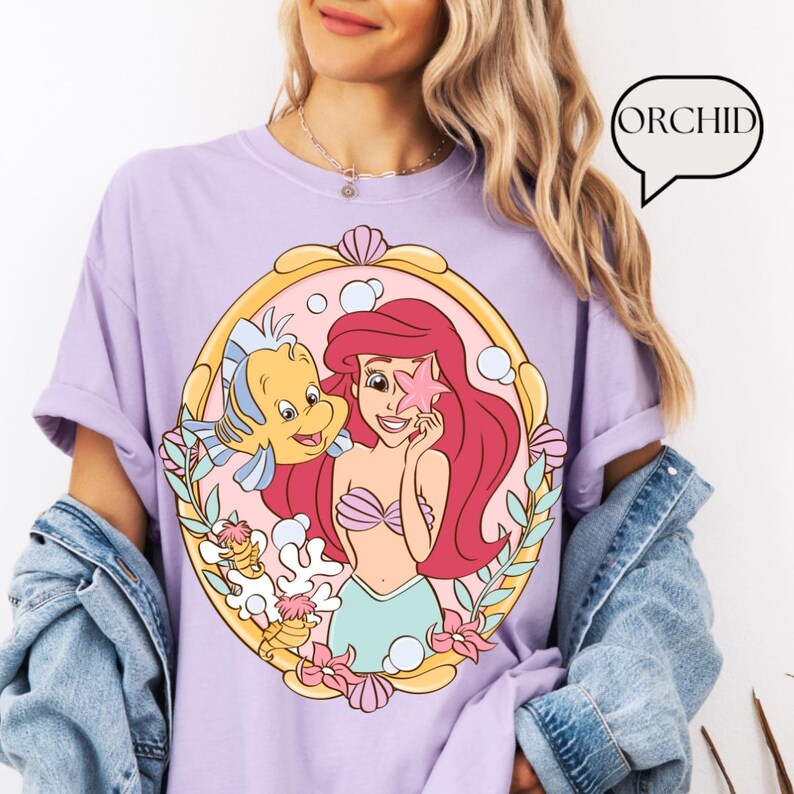 May include: A light purple t-shirt featuring a cartoon illustration of Ariel from The Little Mermaid, holding a starfish with Flounder. The design is framed in a gold oval with pink and green accents. The word "ORCHID" is in a speech bubble.